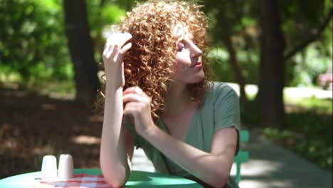 Portrait-of-sensual-curly-ginger-girl-touching-her-red-hair-in-summertime-while-sitting-in-outdoor-park-in-lens-flares.-Enjoying-time-outdoors