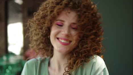 Portrait-of-a-pretty-ginger-girl-with-professional-pink-makeup-smiling.-She-is-having-fun.-Wearing-green-blouse,-twisting-her-head-and-laughing-cheerfuly-to-the-camera,-touching-her-perfect-hairdo.-Outdoors.-Slow-motion