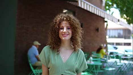 Beautiful-ginger-girl-with-perfect-hairdo---curls-walking-outdoors,-smiling-and-posing-to-the-camera-with-outdoors-cafe-terrace-on-background