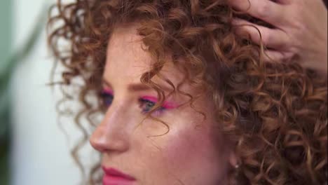 Professional-hair-stylist-finishing-modeling-hairdo-to-young-smilling-girl-customer-with-red-coloured-hair-and-perfect-pink-makeup.-Hairdresser-woman-making-final-touch-to-ginger-long-curly-hair.-Close-up