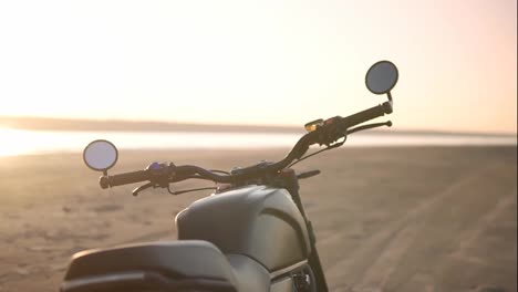 Close-up-footage-of-motorcycle-parking-on-the-ground-close-to-the-water-in-sunset,-sunlight-under-water-on-background