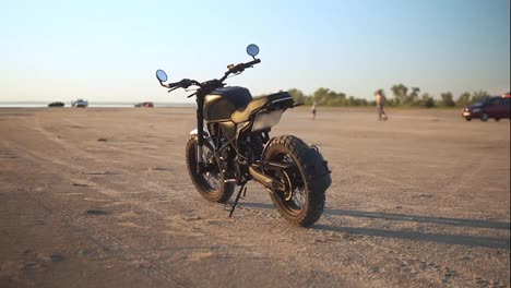 Black-bike-parking-on-the-ground-or-sand-close-to-the-water-in-sunset,-blurred-background-with-people-silhouette
