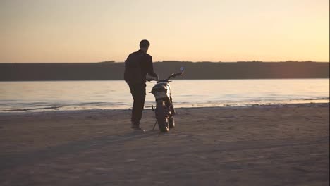 Rare-view-of-a-biker-walking-to-his-bike,-sits-on-it.-The-black-bike-parked-close-to-the-water-with-sunset-view