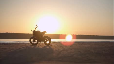 A-motorcycle-parking-on-the-ground-close-to-the-water-in-sunset-with-water-line-on-the-background.-Low-angle-view