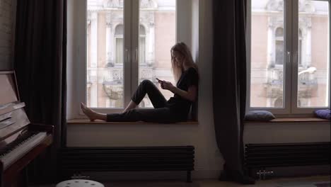 Blonde-woman-sitting-on-window-sill-and-using-smartphone-in-living-room-with-piano