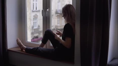 Rare-view-of-a-woman-sitting-on-window-sill-and-using-smartphone-at-home