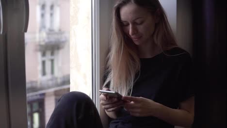 Portrait-of-a-woman-sitting-on-window-sill-and-using-smartphone
