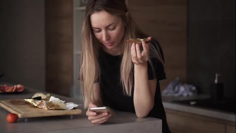 Porträt-Einer-Frau,-Die-Zum-Frühstück-Brot-Mit-Butter-Isst-Und-In-Der-Küche-Ihr-Smartphone-überprüft