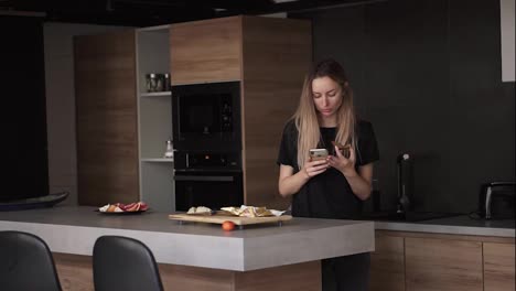 Konzentrierte-Frau,-Die-Zum-Frühstück-Brot-Mit-Butter-Isst-Und-In-Der-Küche-Ihr-Smartphone-überprüft