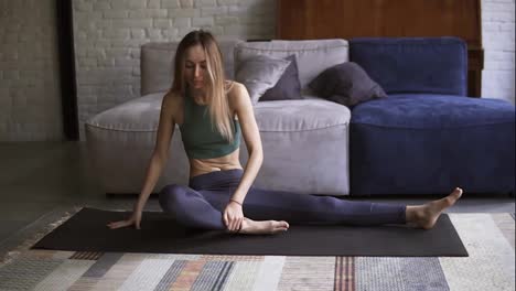 Long-haired-young-woman-is-doing-stretching-exercise-sitting-on-mat-in-living-room