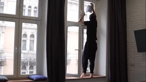 Man-is-cleaning-high-window,-cleans-surface,-uses-a-rag,-standing-on-windowsill