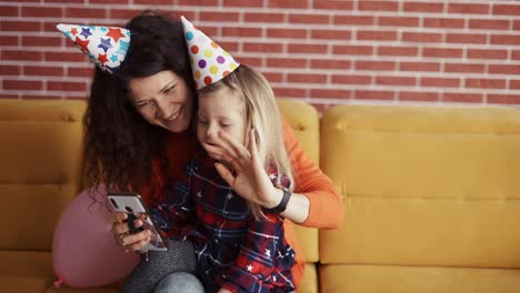 Mother-sitting-with-her-preschool-daughter-in-birthday-caps,-have-videocall-by-mobile