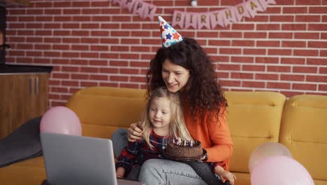 Mother-sitting-with-her-preschool-daughter-in-birthday-caps,-have-videocall-by-laptop
