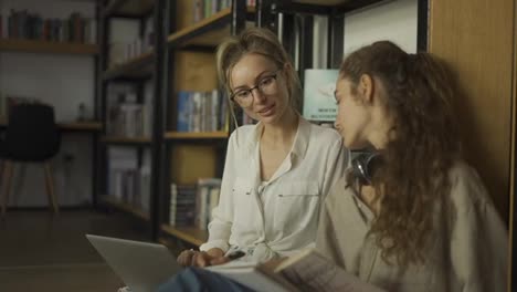 Zwei-Lächelnde-Studentinnen-Lernen-Gemeinsam-Und-Lesen-Bücher-In-Der-Universitätsbibliothek-Auf-Dem-Boden