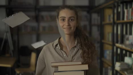 Girl-holding-a-lot-of-books-in-the-library,-paper-sheets-falling-around-her