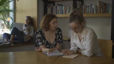 Two-adult-female-students-discussing-new-project-in-library