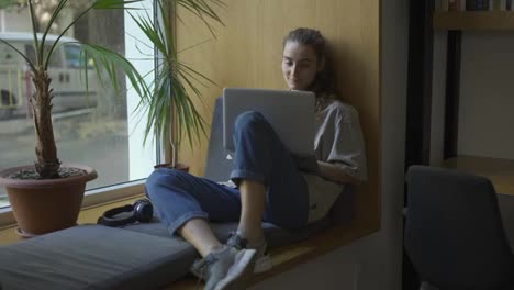Young-attractive-female-freelancer-sitting-on-windowsill-in-cafe-and-working-on-laptop