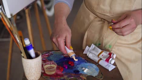 Close-up-footage-caucasian-female-artist-in-apron-squeezing-beige-paint-from-tube-and-putting-on-palette.-Woman,-creative-painter-getting-ready-to-work-over-painting.-Empty-canvas