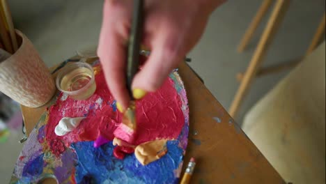 Artist-mixes-pink-paint-on-the-palette-before-painting-a-picture,-painter-at-the-studio,-creator-makes-piece-of-art.-Top-view-of-woman's-hand-with-wide-brush-applying-oil-paint-on-canvas