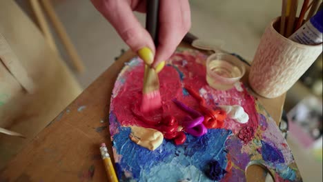 Female-artist-with-thick-paintbrush-is-mixing-different-coloured-oil-paints-on-palette---purple,-red,-yellow.-Close-up-view-of-female-hand-mixing-colours