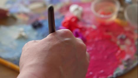 Female-artist-with-thick-paintbrush-is-mixing-different-coloured-oil-paints-on-palette---purple,-red,-yellow.-Close-up-view-of-female-hand-mixing-colours.-Colorful-palette