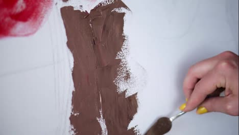 Unrecognizable-artist-paints,-creating-new-art-work-on-the-canvas.-The-artist-paints-using-spatula,-putty-knife-applying-brown-paint-on-white-canvas.-Close-up