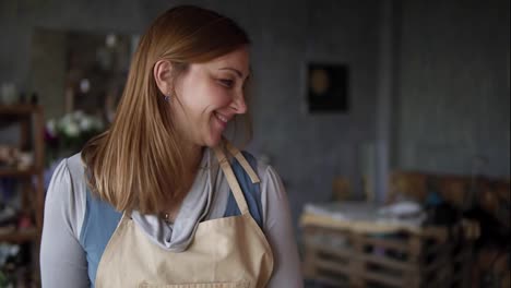 Portrait-of-caucasian-woman-painting-in-arts-studio-using-smear-and-oil-paints.-Long-haired-artist-in-beige-apron,-smiling-while-process