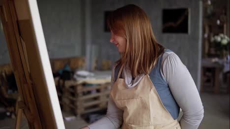 Portrait-of-caucasian-woman-painting-in-arts-studio-using-smear-and-oil-paints.-Long-haired-artist-in-beige-apron,-smiling-while-process,-looking-to-the-camera.-Slow-motion