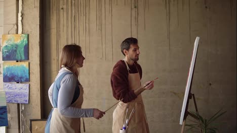 Female-artist-teaching-young-male-student-to-paint-with-oil-paints.-Dark-haired-man-in-beige-apron-drawing-pink-flowers-using-paintbrush.-Workshop-in-art-studio