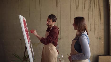 Female-artist-teaching-young-male-student-to-paint-with-oil-paints.-Dark-haired-man-in-beige-apron-drawing-pink-flowers-using-paintbrush.-Workshop-in-art-studio.-Slow-motion
