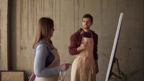 Male-student-studying-to-paint-with-oil-paints.-Dark-haired-man-in-beige-apron-drawing-pink-flowers-using-paintbrush-under-detailed-explanation-by-female-mentor.-Workshop-in-art-studio