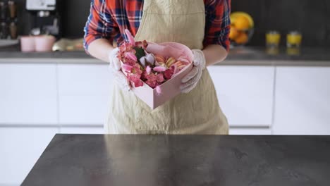Eine-Glückliche-Frau-In-Schürze-Präsentiert-Eine-Schachtel-Mit-Makronen-Und-Mit-Blumen-Verzierten-Keksen