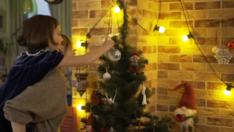 Mother-holding-girl-on-back,-hanging-together-Christmas-tree-toys-on-top-of-Christmas-tree