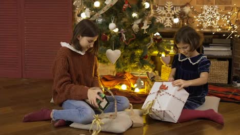 Zwei-Aufgeregte-Kleine-Mädchen-Schütteln-Eine-Geschenkbox-Unter-Dem-Weihnachtsbaum,-Der-Auf-Dem-Boden-Sitzt