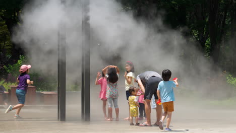 Group-of-Children-Have-Fun-Cooling-Down-On-Hot-Summer-Day-Under-Misting-Micro-Flow-Drip-Cooling-System-in-Public-Park,-Sprinking-Cold-Aero-Mist---Yangjae-Citizen-Forest
