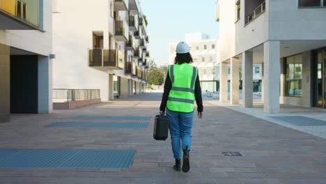 Female-Engineer-Wearing-High-Visibility-Vest-Carrying-Small-Case-Along-Pavement-In-Urban-Setting