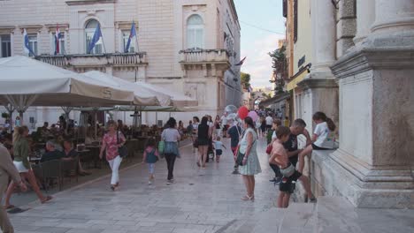 Zadar-old-town-with-summer-crowds,-pedestrians,-and-a-women-selling-balloons,-and-a-cafe