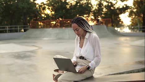 Young-elegant-caucasian-woman-with-dreadlocks-in-white-blouse-sitting-in-summer-park.-Happy-freelancer-typing-on-laptop-outdoors