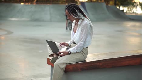 Side-view-of-beautiful,-elegant-girl-with-black-and-white-dreadlocks-sitting-on-parapet-in-local-skatepark-working-on-laptop