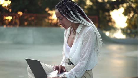 Side-view-of-beautiful,-elegant-girl-with-black-and-white-dreadlocks-sitting-on-parapet-in-local-skatepark-working-on-laptop