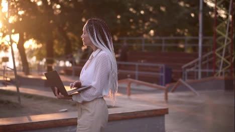 Lovely-woman-with-black-and-white-dreadlocks-hair-walking-by-the-street-with-open-silver-laptop,-typing.-Lens-flares-and-trees