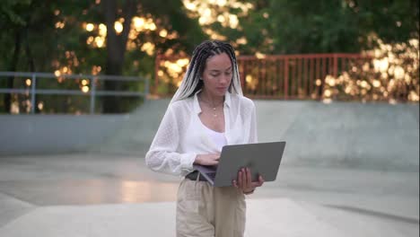 Elegant,-young-woman-with-black-and-white-dreadlocks-hair-walking-by-park-or-skatepark-with-open-silver-laptop,-typing.-Wearing