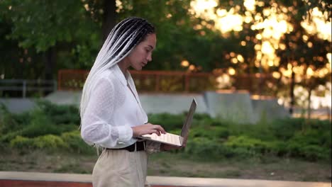 Lovely-woman-with-black-and-white-dreadlocks-hair-walking-by-the-street-with-open-silver-laptop,-typing.-Lens-flares-and-trees