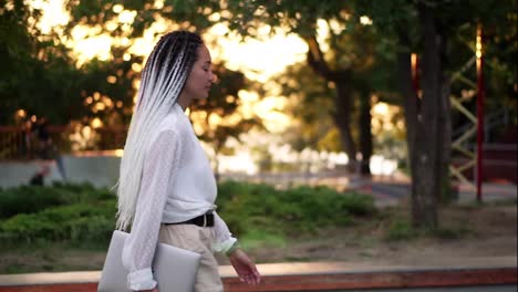 Confident-walk-of-and-elegant-girl-in-white-blouse-and-pants---young-girl-walks-through-the-park-with-a-laptop-in-her-hand.-Side