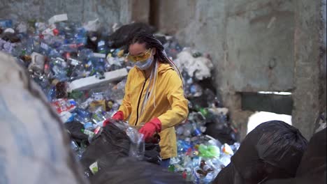 Stylish-woman-in-yellow-jacket,-protective-glasses-and-gloves-sorting-plastic-bottles-from-black-bags.-Huge-pile-of-used-plastic-bottles-on-background