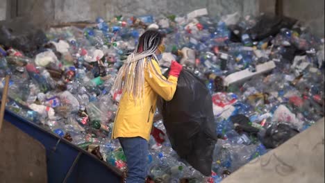 Woman-worker-in-yellow-jacket-and-protecting-glasses-unpacking-black-sucks-with-used-plastic-bottles-at-modern-recycling-plant.-Throwing-bottles-from-big-sack-on-pile-in-recycling-factory