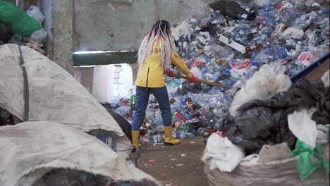 A-rare-view-of-a-girl-with-dreadlocks,-in-yellow-boots,-scoops-used-bottles-with-a-large-duck-shovel-at-a-plastic-recycling-factory.-Huge-pile-of-bottles-on-background