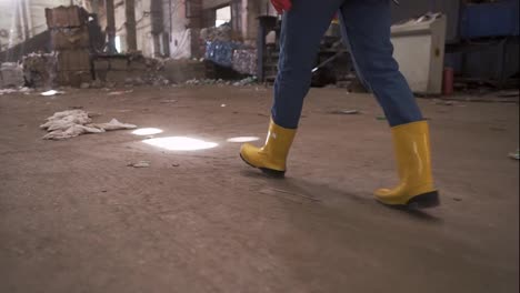 Unrecognizable-woman-in-protective-working-clothes----rubber-gloves-and-boots-walking-by-waste-recycling-factory,-huge-area-with-used-bottles,-recycling-plant