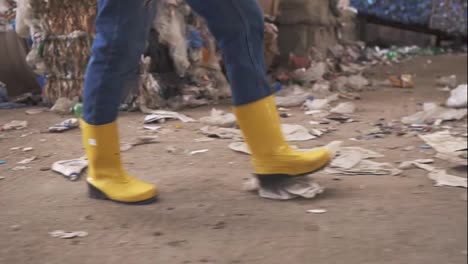 Unrecognizable-woman-in-protective-working-clothes---yellow-boots-walking-by-waste-recycling-factory,-huge-area-with-used-bottles-and-garbage.-Close-up-footage
