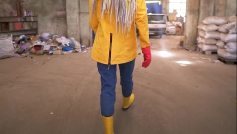 Tracking-rare-view-footage-of-a-girl-in-protective-working-clothes---walking-by-waste-recycling-factory,-huge-area-with-used-bottles,-different-waste-and-white-truck-on-background.-Close-up